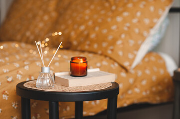 Wall Mural - Home liquid perfume in glass bottle with bamboo sticks and burning candle on paper book staying on rattan table close up in bedroom. Cozy atmosphere.