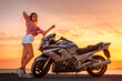 Pretty young woman standing posing on a motorcycle. Golden sunset and ocean on the background. Freedom, World Motorcyclist Day and moto trips