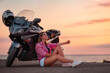 Smiling young woman sitting and posing near motorcycle. Golden sunset and motorbike on the background. Copy space. Concept of World Motorcyclist Day and moto travel