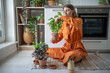 Interested woman takes care of home seedlings sits on kitchen floor, cuts leaves of green basil herbs. Female gardener pays attention to care of houseplants, tends, prunes excess and harvests crop.