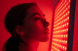 © Darya - Side profile of a young woman bathed in the crimson glow of a red light therapy panel, exuding a sense of contemplation and wellness.