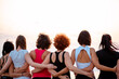 Back view of group of fit women in sportswear stands in row and hugs each other. Light sunset sky in background. Copy space. Concept of girl power and sports competition