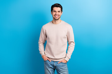 Wall Mural - Portrait of funny satisfied man with brunet hairstyle dressed beige sweatshirt standing arms in pockets isolated on blue color background
