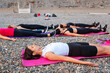 Yoga class at sea coast. Side view of group of Caucasian women lies in shavasana on sports mats on pebble beach. Meditation outdoor training