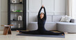 A woman does yoga at home, performs the exercise Samakonasana, transverse twine, trains in a black sports overalls on the mat, healthy lifestyle concept
