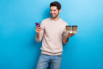 Aufkleber - Portrait of positive nice handsome man look at smartphone holding two cups of coffee read notification isolated on blue color background