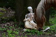 Abschied im Wald. Symbolisches Bild vom Abschied. Menschliches Skelett sitzt im Wald und hält skelettierten Pferdeschädel während ein schönes Pferd anbei grast