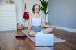 Portrait of young and relaxed woman using laptop and wireless headphones while meditating, practice yoga at home on rubber mat, sitting in asana