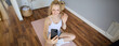 Portrait of fit, sporty young fitness instructor, woman gives online workout sessions to clients, waves hand at smartphone, sits on yoga mat and shows exercises