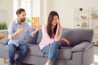 Angry husband shouting in quarrel on his unhappy upset wife sitting on sofa at home. Young couple quarreling and arguing in living room. Marriage problems, relationship and divorce concept.