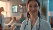 A confident female doctor in a crisp white coat and stethoscope smiles warmly in a bright hospital waiting room, framed by a backdrop of cheerful patients and soft ambient light.