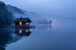 A small cabin sits on a dock in front of a lake. The water is calm and the sky is cloudy