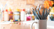 White mug with colored pencils in the foreground on a wooden table with art supplies in the background