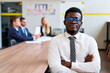 Confident African corporate professional in formal workwear sits at meeting table, focus on leadership with diverse team blurred in background. Inclusive modern office setting, busy workday.