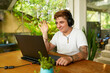 Smiling Gen Z Transgender pro in casual attire enjoys video call on laptop in coworking space. Wave hello, engaged in remote work surrounded by green plants, embracing digital nomad lifestyle.