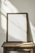 Close-up of an empty vertical space with a wooden frame standing on an old wooden table leaning against a whitewashed wall, illuminated by sunlight.