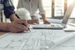 A closeup shows two architects working on blueprints, with one holding a white hard hat and the other drawing lines onto paper using pencils Generative AI