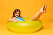 Happy funny little girl dressed in a blue swimsuit lying on an inflatable color ring on yellow background. Summer vacation concept. Beach time. Copy space for ad.
