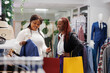 Smiling women shoppers holding jacket on hanger and discussing apparel style in clothing store. Cheerful african american friends carrying purchase packages while choosing formal blazer in boutique