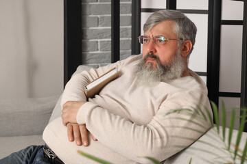 Poster - Portrait of sad senior man with book sitting on sofa in living room