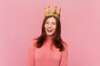Happy confident smiling woman with brown hair standing in gold crown, looking at camera with positive expression, laughing, wearing rose turtleneck. Indoor studio shot isolated on pink background