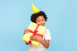 © khosrork - Portrait of cute little boy with curly hair wearing yellow party cone standing embracing his birthday present enjoying with closed eyes. Indoor studio shot isolated on blue background.
