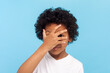 © khosrork - Portrait of shy curious little boy with curly hair looking at camera trough his fingers spying sees something shameful. Indoor studio shot isolated on blue background.