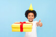© khosrork - Portrait of joyful little boy with curly hair wearing yellow party cone holding wrapped yellow present box showing thumb up. Indoor studio shot isolated on blue background.