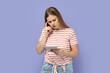Portrait of thoughtful attentive beautiful blond woman wearing striped T-shirt holding and working on tablet reading something with pensive expression. Indoor studio shot isolated on purple background