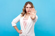 Portrait of strict bossy blond woman with wavy hair showing palm stop gesture prohibition sign looking at camera with serious face, wearing white shirt. Indoor studio shot isolated on blue background.