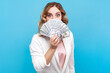 Portrait of shocked woman with wavy hair covering half of face with fan of money standing with her salary astonished expression, wearing white shirt. Indoor studio shot isolated on blue background.
