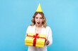 Portrait of shocked surprised blond birthday woman with wavy hair holding yellow wrapped present box celebrating holiday, wearing white shirt. Indoor studio shot isolated on blue background.