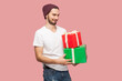 Portrait of cunning smiling bearded man in white T-shirt and beany hat standing holding present boxes and looking at camera with sly expression. Indoor studio shot isolated on pink background.