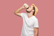 Side view portrait of tired bearded man in white T-shirt and hat standing holding disposable cup of coffee, drinking hot beverage. Indoor studio shot isolated on pink background.
