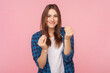 Portrait of smiling sly brown haired woman standing showing money gesture looking at camera reward, wearing checkered shirt. Indoor studio shot isolated on pink background.