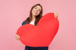 Portrait of smiling good looking brown haired woman holding big red heart looking at camera with positive expression, wearing checkered shirt. Indoor studio shot isolated on pink background.