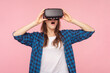 Portrait of shocked surprised woman in vr headset, playing virtual reality game with open mouth, innovative technology, wearing checkered shirt. Indoor studio shot isolated on pink background.