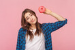 Portrait of confused uncertain brown haired woman holding delicious sugary donut looking at camera with puzzled expression, wearing checkered shirt. Indoor studio shot isolated on pink background.