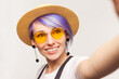 Portrait of smiling delighted woman blogger with violet hair in sunglasses and hat making point of view photo livestream. Indoor studio shot isolated on white background.