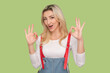 Portrait of excited positive adult blond woman standing looking at camera, saying everything is ok, showing okay gesture, wearing denim overalls. Indoor studio shot isolated on light green background
