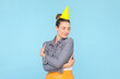 Portrait of cute heartwarming woman with bun hairstyle wearing denim jacket and party cone embracing herself celebrating birthday. Indoor studio shot isolated on light blue background.