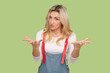 What do you want? Portrait of unhappy sad adult blond woman looking at camera with negative expression, being frustrated, wearing denim overalls. Indoor studio shot isolated on light green background
