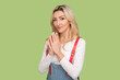 Portrait of envy cunning beautiful adult blond woman presses fingers together, planning sly prank, looking at camera, wearing denim overalls. Indoor studio shot isolated on light green background