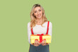 Portrait of satisfied pleased pretty adult blond woman holding present box in hands, congratulating with birthday, wearing denim overalls. Indoor studio shot isolated on light green background