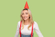 Portrait of charming delighted adult blond woman in party cone looking smiling at camera, being in good festive mood, wearing denim overalls. Indoor studio shot isolated on light green background