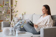 Young woman reading magazine on sofa in stylish living room with vases of blooming branches
