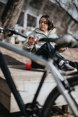 Wall Mural - Relaxed young girl sitting in a park, checking her phone with a bicycle and a drink beside her on a sunny day.