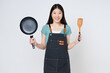 Young woman wearing kitchen apron cooking and holding pan and spatula isolated on white background.