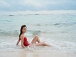 © SHOTPRIME STUDIO - Woman with a beautiful tan tourist in a red swimsuit sitting on the sand on the beach in the ocean in the waves pensive