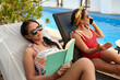 © DragonImages - Teenage girl reading book when sunbathing by pool with her mother